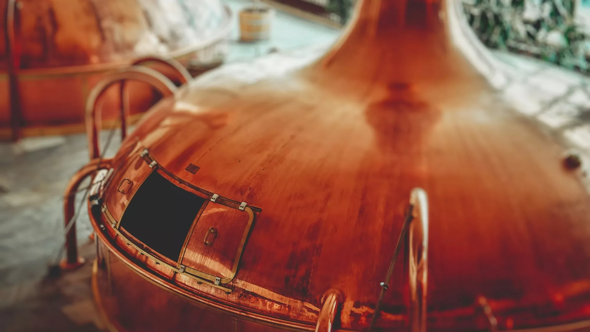 Großer kupferner Braukessel mit offener Luke, umgeben von Metallgeländern, in einer Brauerei mit weichem natürlichem Licht und unscharfem Hintergrund.