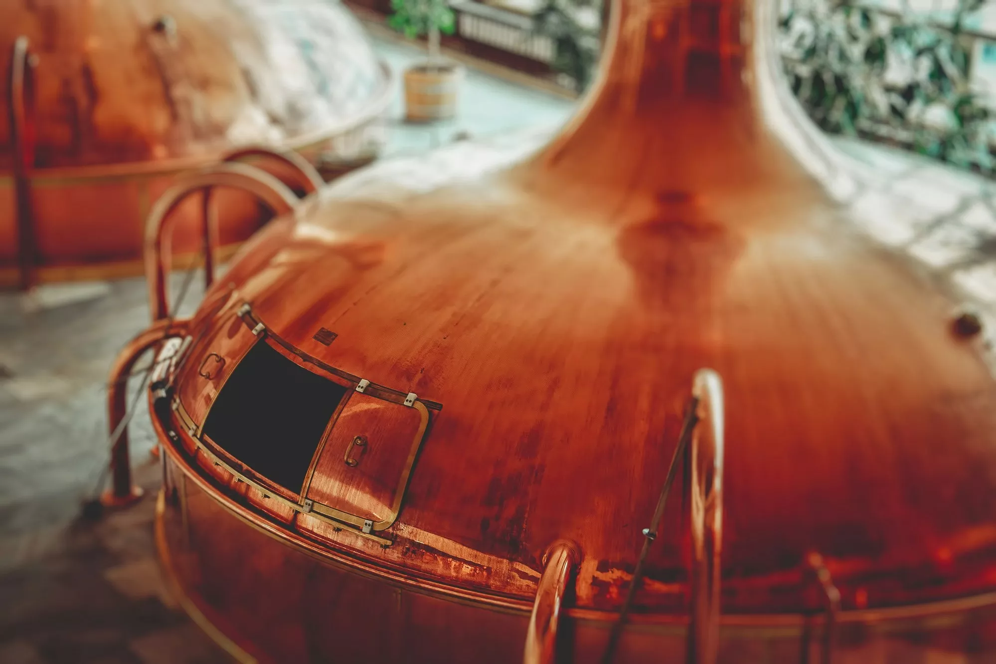 Großer kupferner Braukessel mit offener Luke, umgeben von Metallgeländern, in einer Brauerei mit weichem natürlichem Licht und unscharfem Hintergrund.
