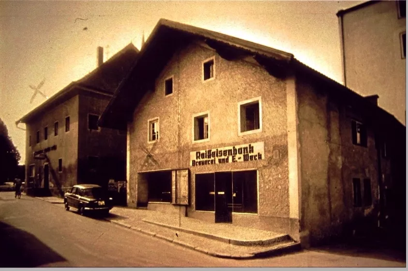 Sepiafarbenes Foto einer Straßenecke mit einem alten Gebäude, an dem ein Schild mit der Aufschrift "Raiffaisenbank Brauerei und E. Werk" angebracht ist. Ein Oldtimer ist am Straßenrand geparkt, und im Hintergrund ist schwach eine Windmühle zu erkennen.