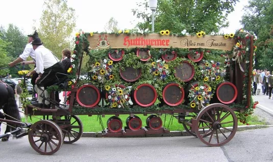 Ein pferdegezogener Festwagen zum 400-jährigen Jubiläum Festzug ist mit Blumen und Grünzeug geschmückt, zeigt mehrere Bierfässer und ein Schild mit der Aufschrift "Hutthurmer - Alles vom Feinsten". Vorne sitzen zwei Personen in Tracht.